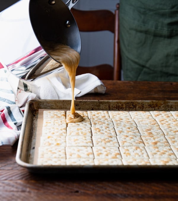 A woman pours liquid toffee over a layer of saltine crackers laid out on a rimmed baking sheet.