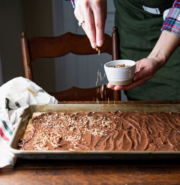 A woman sprinkles chopped pecans over the entire top of a sheet pan of Christmas crack toffee.