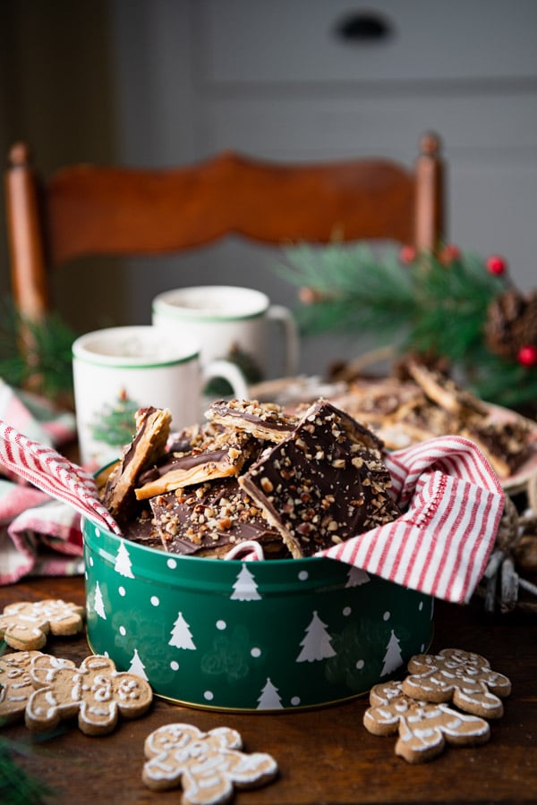A side-angle image of a full tin of saltine cracker toffee candy sitting on a tabletop, surrounded by mini gingerbread cookies and mugs of hot cocoa.