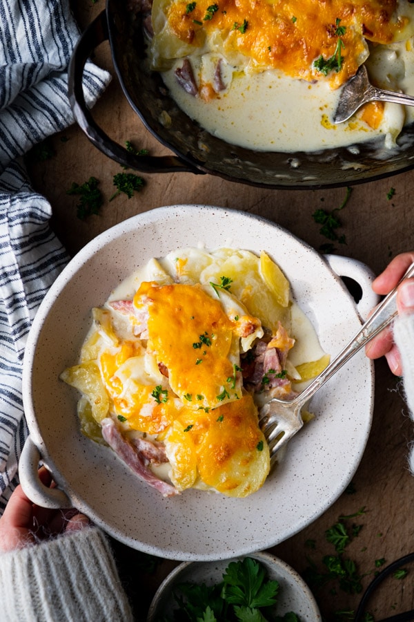 Overhead shot of hands holding a fork to eat cheesy scalloped potatoes and ham