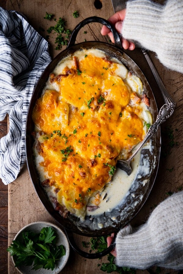 Hands holding a pan of cheesy scalloped potatoes with ham on a wooden board