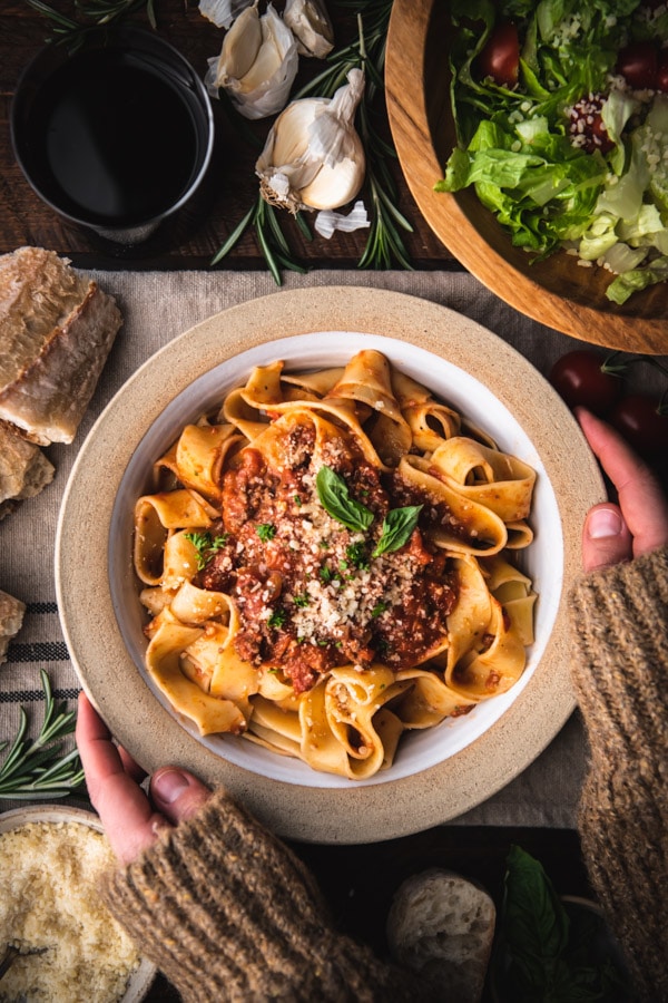Hands holding a bowl of the best bolognese recipe garnished with herbs and Parmesan cheese.