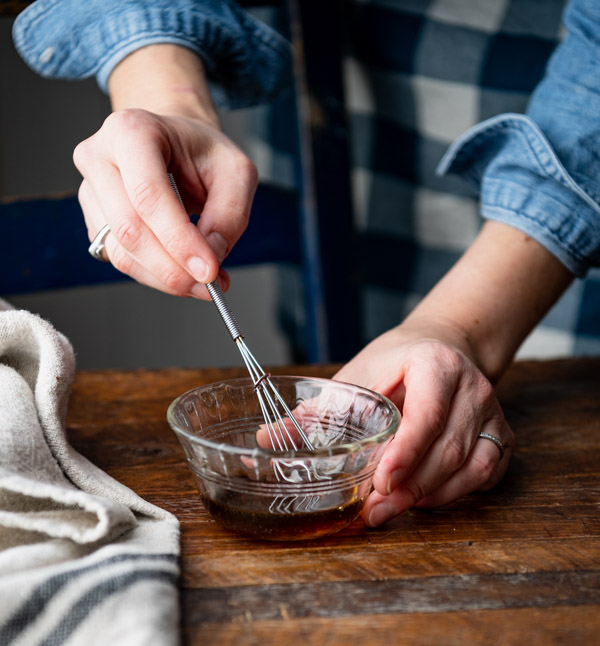 A woman uses a small whisk to mix together a honey maple glaze in a small glass bowl. 