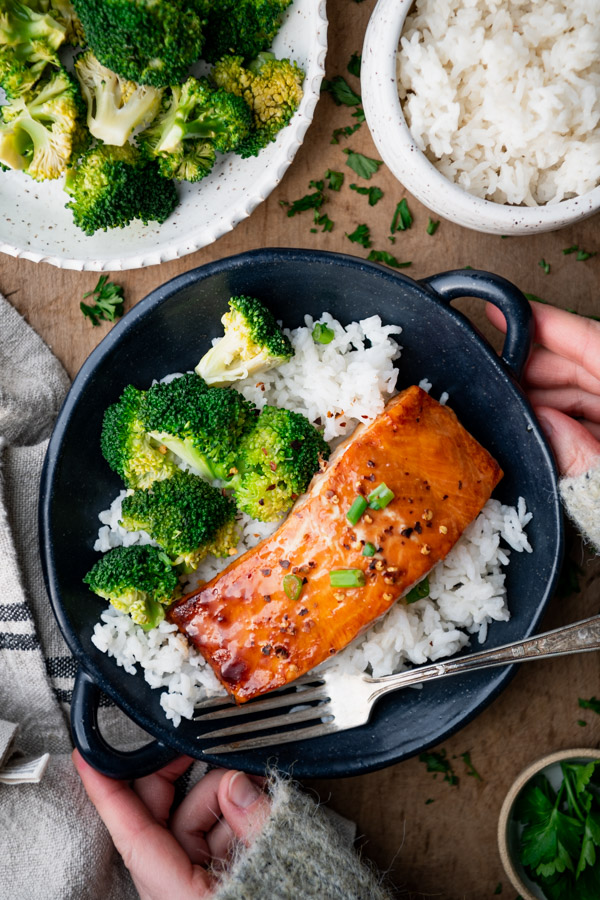 An overhead image of a large bowl with one fillet of maple glazed baked salmon served over white rice with broccoli. A large plate of cooked broccoli and a bowl of white rice sit on the table next to the plate.