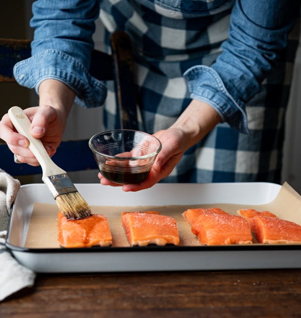 A woman brushes soy sauce over four raw salmon fillets lined up on a parchment paper lined baking sheet.
