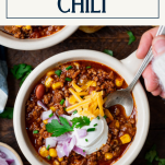 Overhead shot of hands eating a bowl of beef chili with text title box at top.