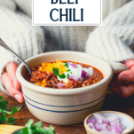 Side shot of hands holding a bowl of the best beef chili recipe with text title overlay.