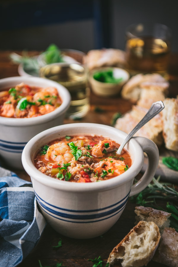 Two mugs of slow cooker cioppino with herbs, wine, and bread in the background
