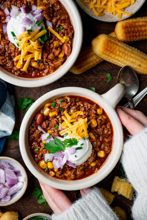 Hands holding a bowl of chili con carne with cheese and sour cream on top.