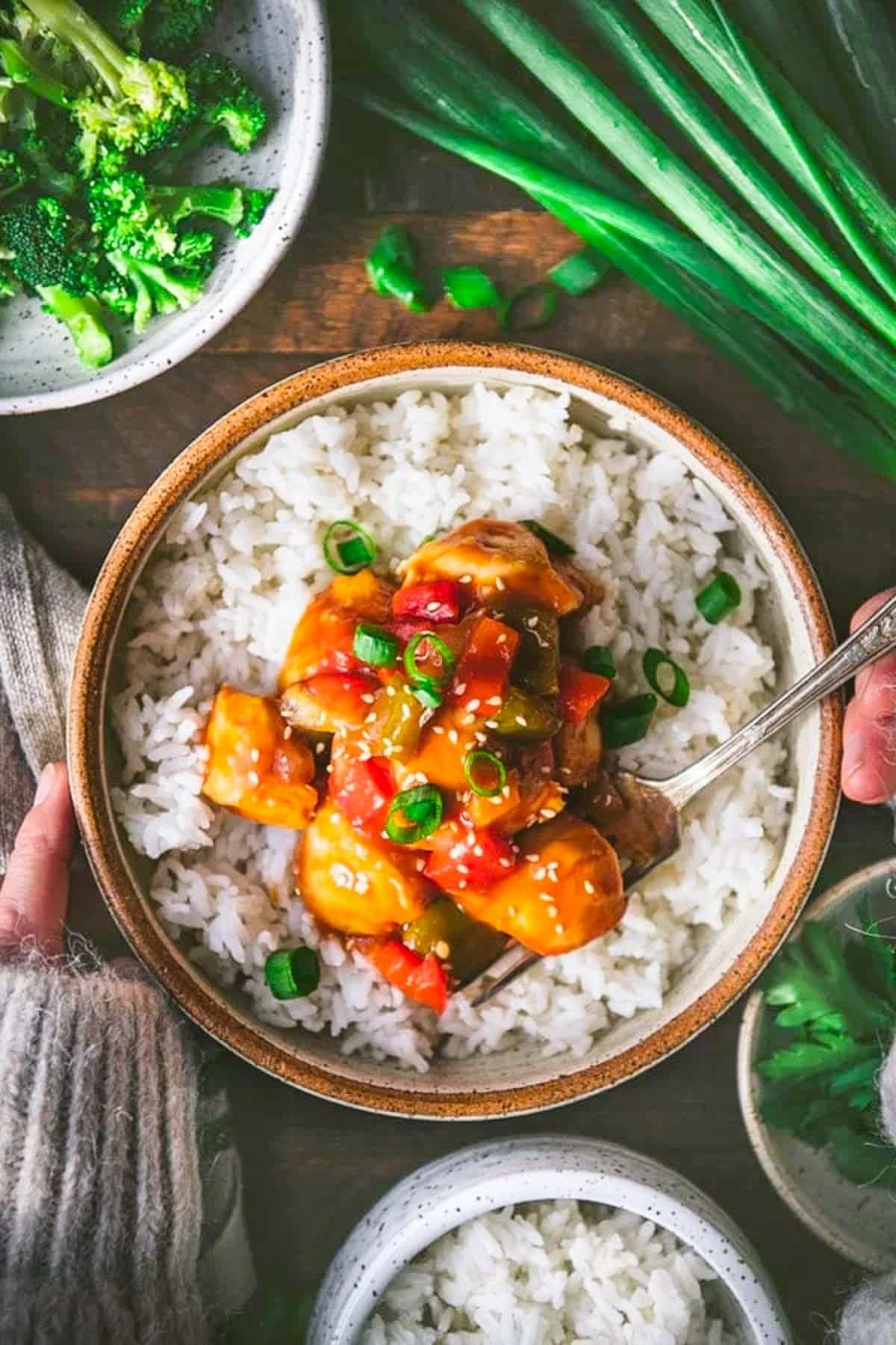 Overhead shot of hands eating a bowl of baked sweet and sour chicken with rice on a wooden table.