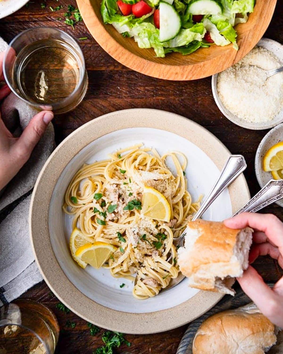 Hands holding a piece of French bread alongside a bowl of linguine with clam sauce.