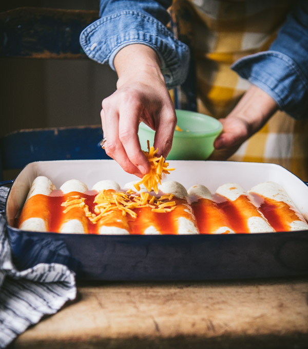 A woman sprinkles shredded cheddar cheese over the top of unbaked beef enchiladas, which are covered in red enchilada sauce.