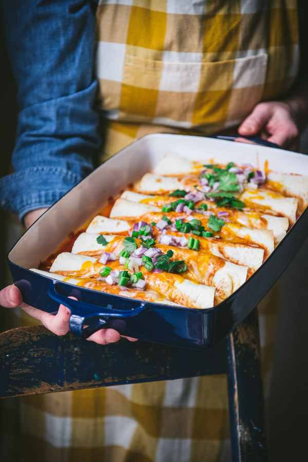 A woman holds a blue and white casserole dish filled with homemade beef and cheese enchiladas, garnished with diced red and green onions.