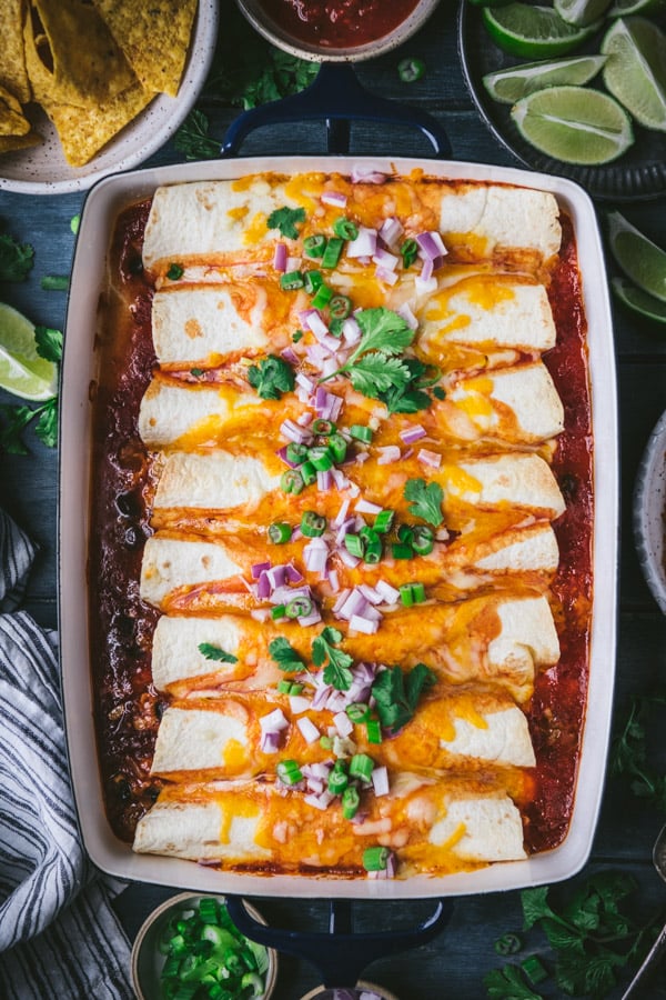 An overhead image of 8 beef and cheese tortillas in a casserole dish with enchilada sauce, shredded cheese, and red and green onions for garnish.