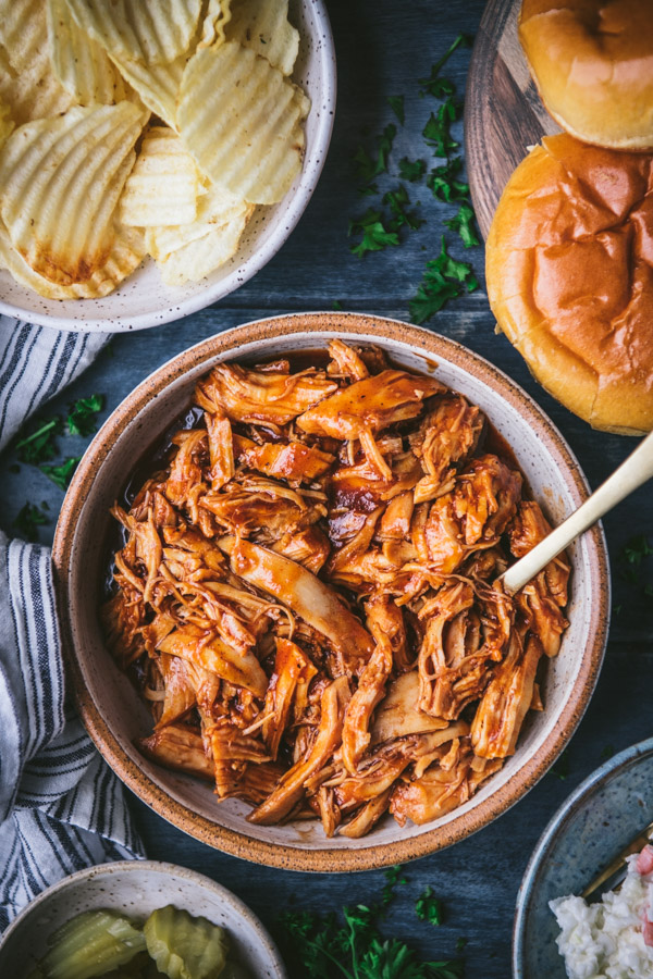 An overhead image of BBQ pulled chicken in a larger bowl, surrounded by bowls of pickles, coleslaw, potato chips, and brioche hamburger buns on a platter.