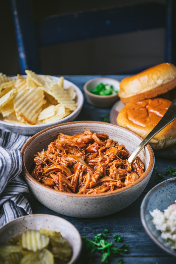 A bowl full of BBQ pulled chicken set on a tablescape with bowlfuls of other ingredients - pickle slices, potato chips, hamburger buns, and creamy coleslaw.