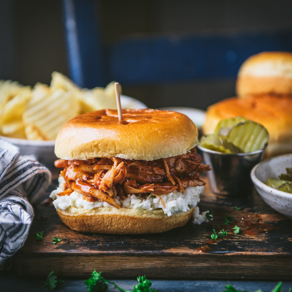 A juicy BBQ pulled chicken sandwich sitting on a wooden cutting board, surrounded by plates of sides -- pickles, coleslaw, and potato chips.