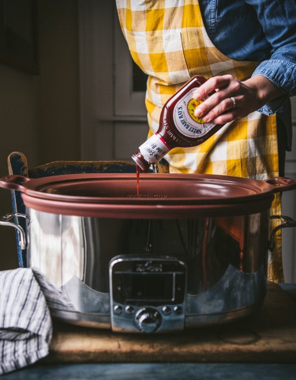 A woman pours a bottle of Sweet Baby Ray's BBQ sauce into a large crockpot.