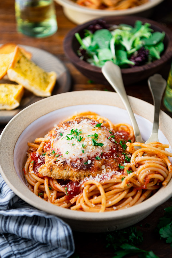 Close up side shot of chicken parmesan with pasta in a bowl on a wooden dinner table.