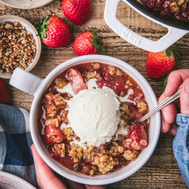 Overhead shot of hands eating a bowl of strawberry crisp with vanilla ice cream on top