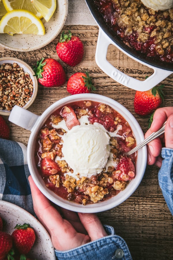Overhead shot of hands eating a bowl of strawberry crisp with vanilla ice cream on top