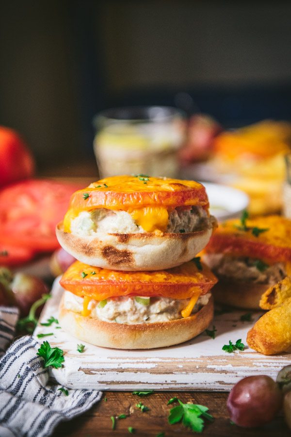 Two tuna melts stacked on top of each other on a wooden cutting board. The tuna melts are made on English muffins, topped with tomato slices and melted cheese.