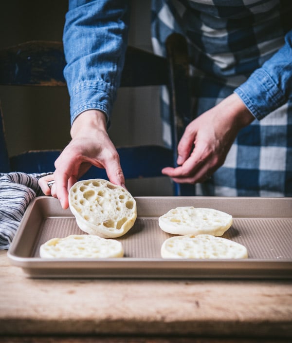 A woman places two open faced English muffins on a baking sheet, preparing them to be toasted.