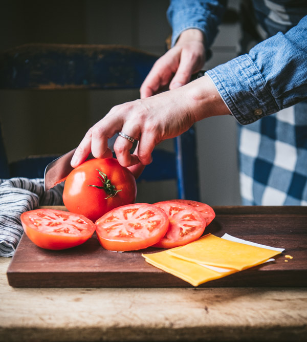 A woman slices thick slices of tomato on a cutting board, next to slices of cheddar cheese.