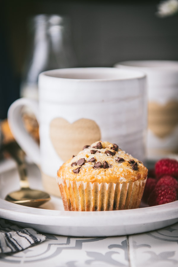Side shot of a chocolate chip muffin on a white plate with a mug of coffee and fresh berries