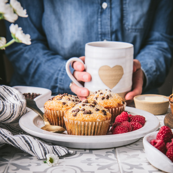 Front shot of a square image of a plate of the best chocolate chip muffins with fresh berries and a person holding a mug of coffee