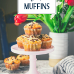 Cake stand with chocolate chip muffins and flowers in the background and text title overlay