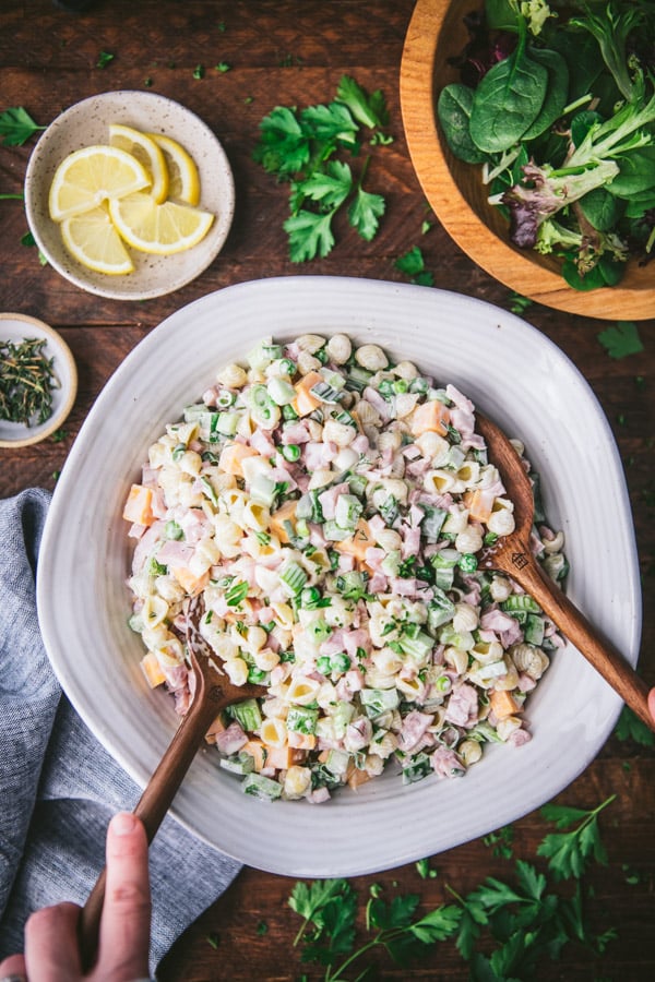 Overhead shot of hands serving ham pasta salad with two wooden spoons