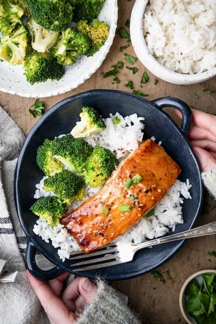 Overhead shot of hands holding a bowl of honey maple glazed baked salmon on a wooden table.