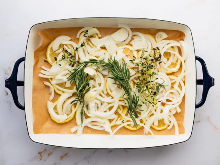 Lemon slices, sliced onions, fresh herbs, and garlic in the bottom of a baking dish.