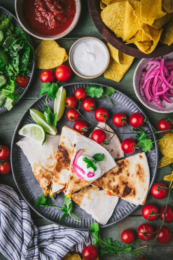 Overhead shot of a plate of chicken quesadillas with tomatoes, limes, cilantro, and pickled red onions.