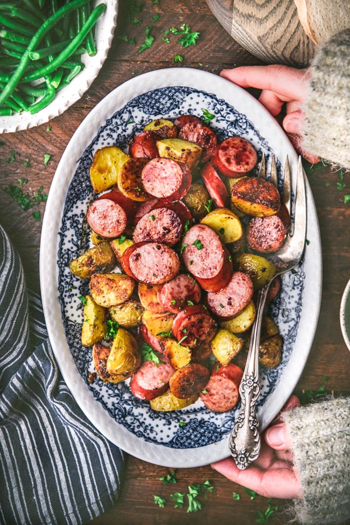 Overhead shot of hands holding a tray of sheet pan sausage and potatoes on a wooden table.