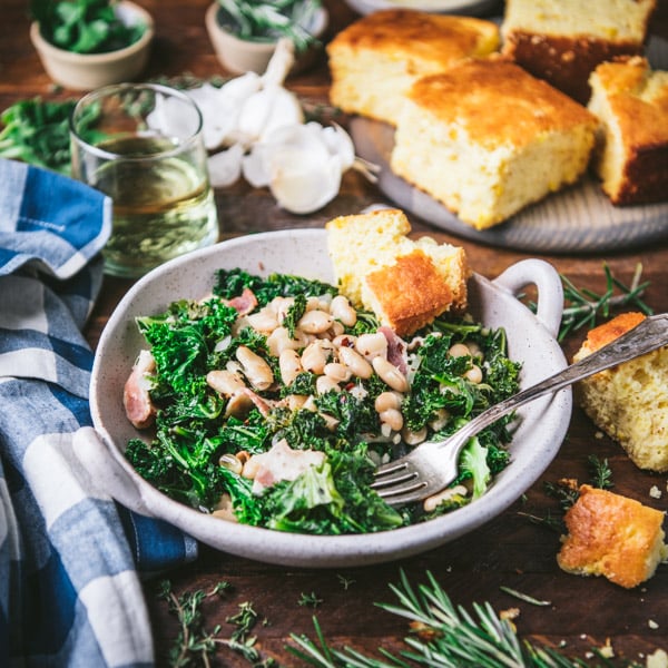 Square side shot of greens and beans in a bowl on a wooden table with a side of cornbread.