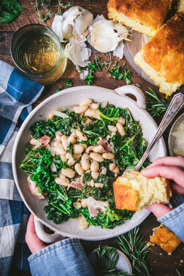 Overhead shot of hands eating a bowl of beans and greens with cornbread