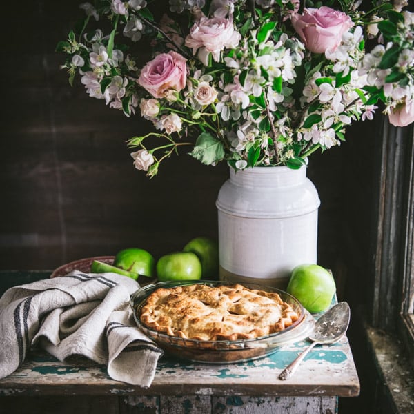 Apple pie in a glass dish on a wooden chest with flowers in the background