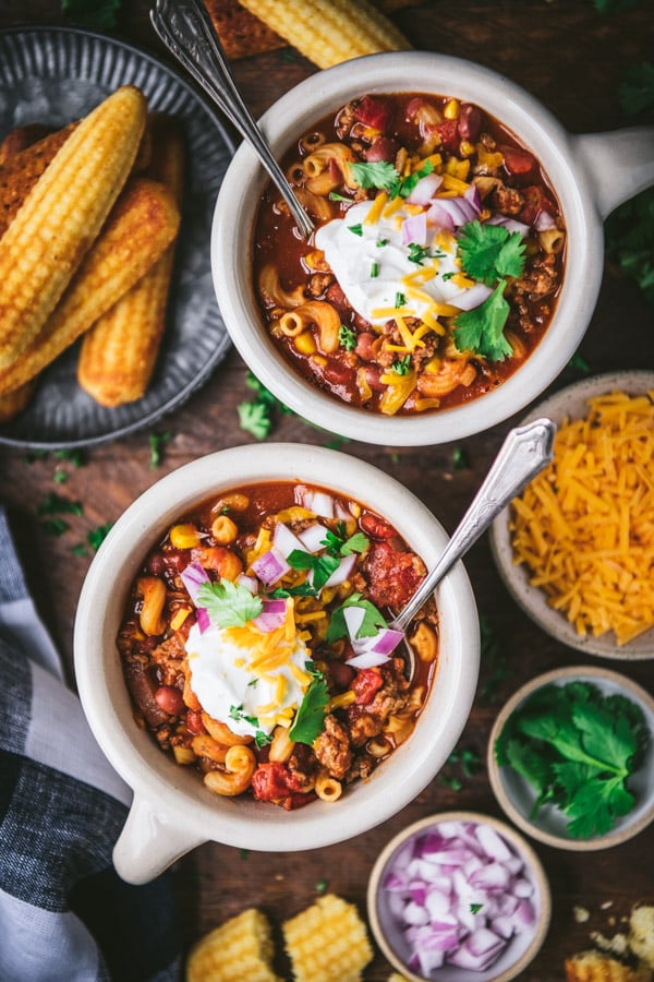 Two bowls of chili mac served on a wooden dinner table with a side of cornbread