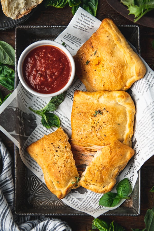 Pizza pockets in a rectangular metal tray with fresh herbs and a bowl of marinara sauce for dipping
