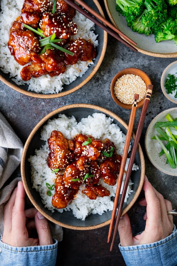 Overhead shot of hands holding a bowl of homemade sesame chicken with rice