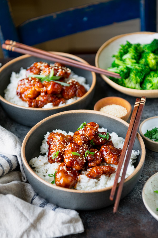 Side shot of two bowls of sesame chicken with rice and a side of broccoli