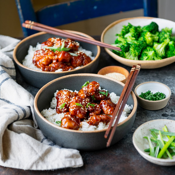 Square image of a side shot of two bowls of sesame chicken on a dinner table