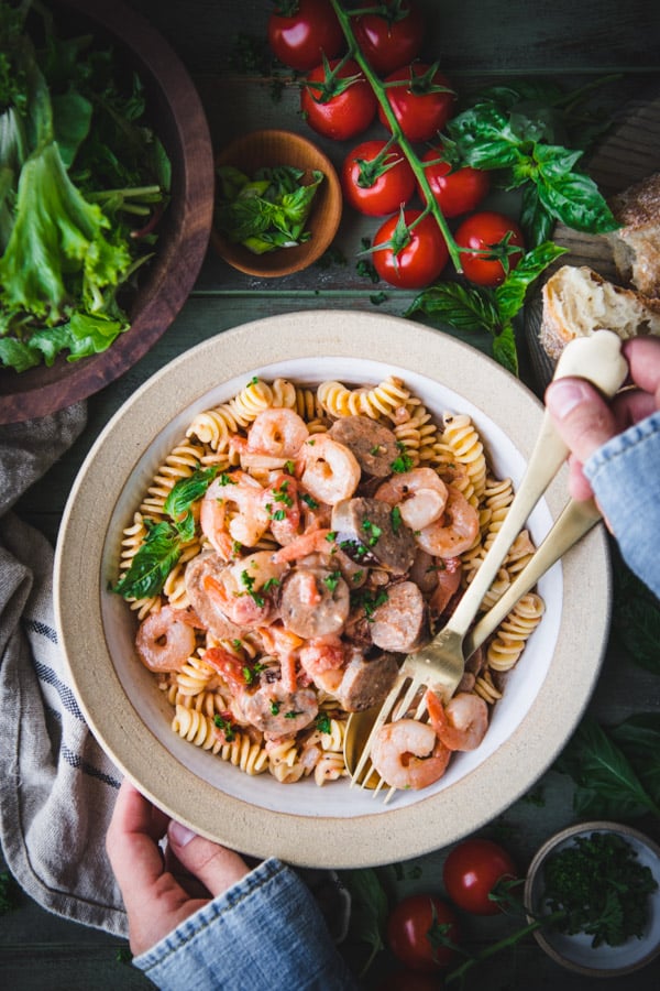 Hands eating a bowl of sausage and shrimp pasta with gold fork and spoon