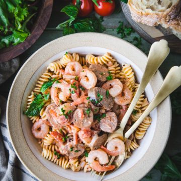 Close overhead shot of a bowl of shrimp sausage pasta on a green table with salad and bread on the side