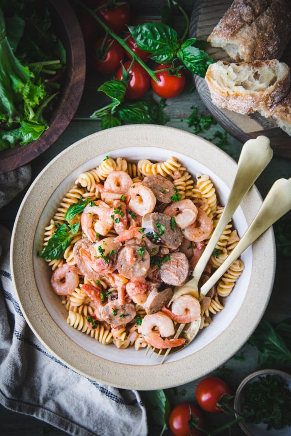 Close overhead shot of a bowl of shrimp sausage pasta on a green table with salad and bread on the side