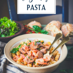 Side shot of a bowl of shrimp and sausage pasta on a table with bread and salad and text title overlay