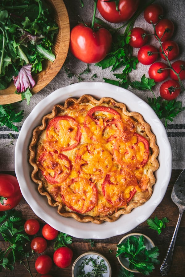 Overhead shot of a tomato pie in a white dish on a table with fresh tomatoes and salad