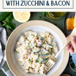Hands eating a bowl of cheesy pasta with zucchini and bacon and text title box at top